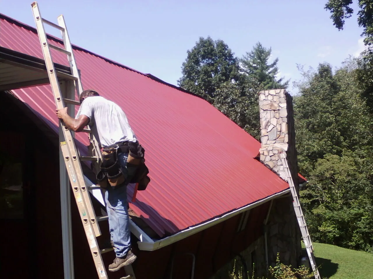 Roofer on a ladder beside a red metal roof and stone chimney — Soffit & Fascia Repair by Roofing Brothers in New Boston